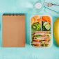School lunch consisting of sandwich, fresh fruits and vegetables sitting next to notepad.