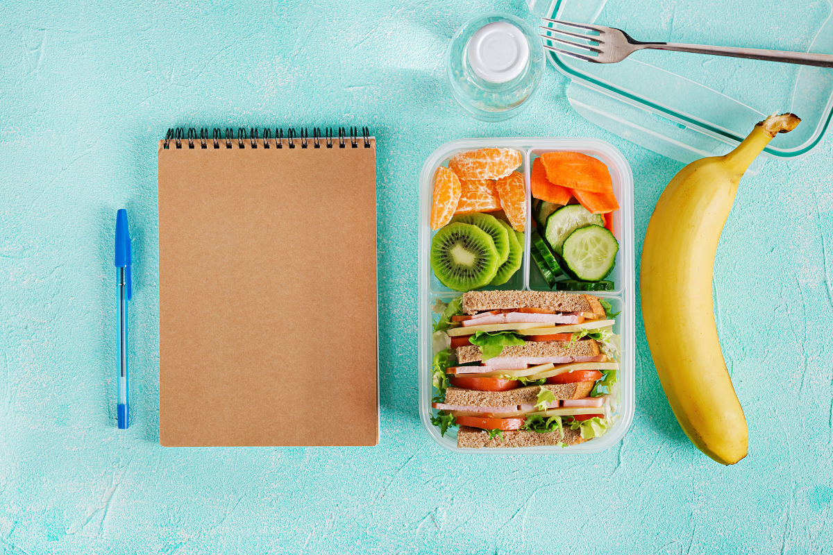 School lunch consisting of sandwich, fresh fruits and vegetables sitting next to notepad.